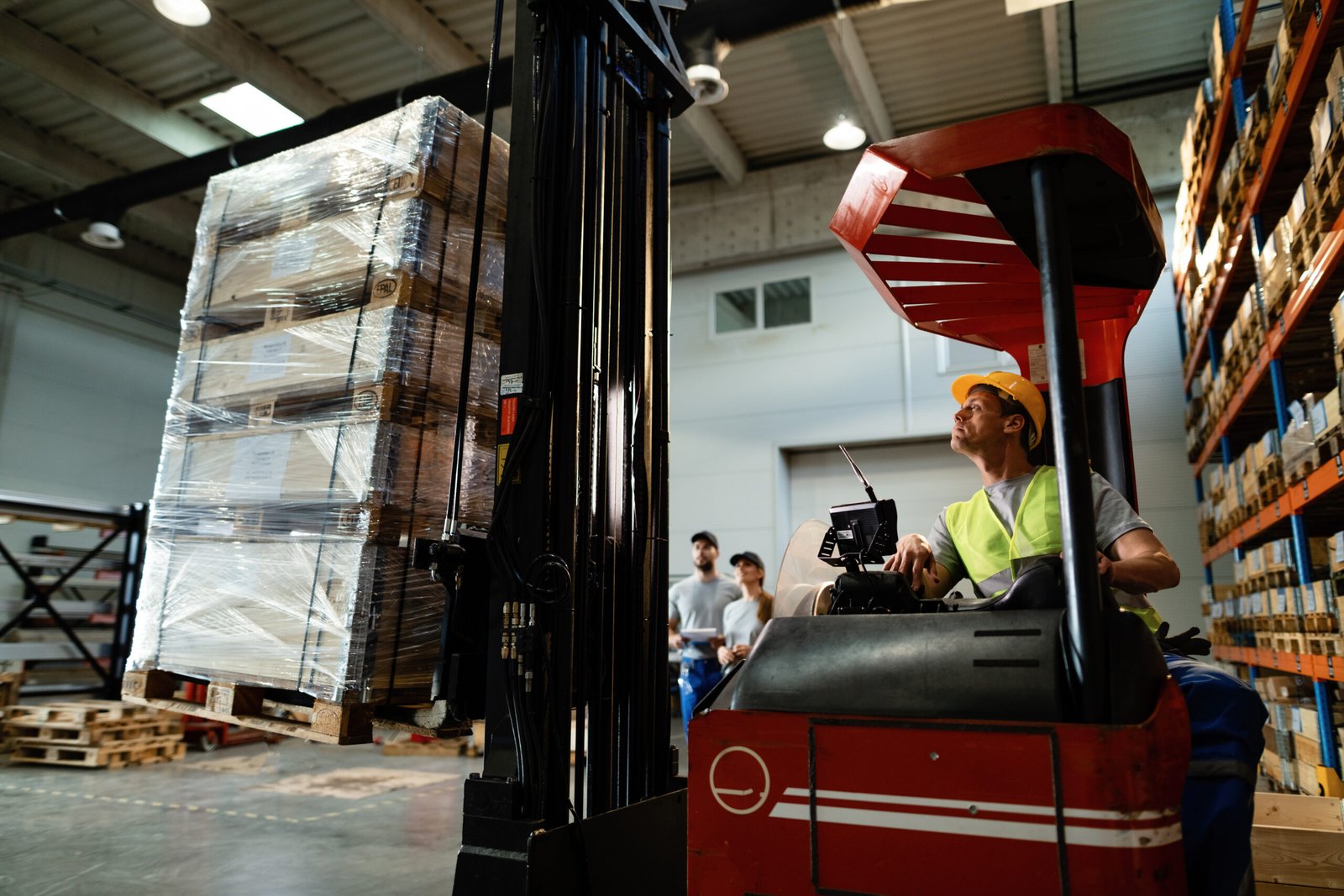 Forklift operator loading cargo while working in a warehouse. His colleagues are in the background.